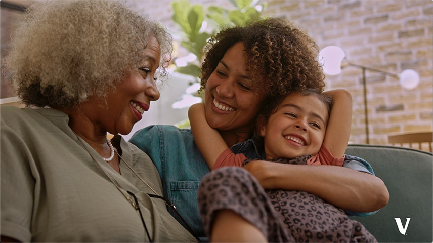 A mother and grandmother are smiling at each other and seated next to each other on a couch
